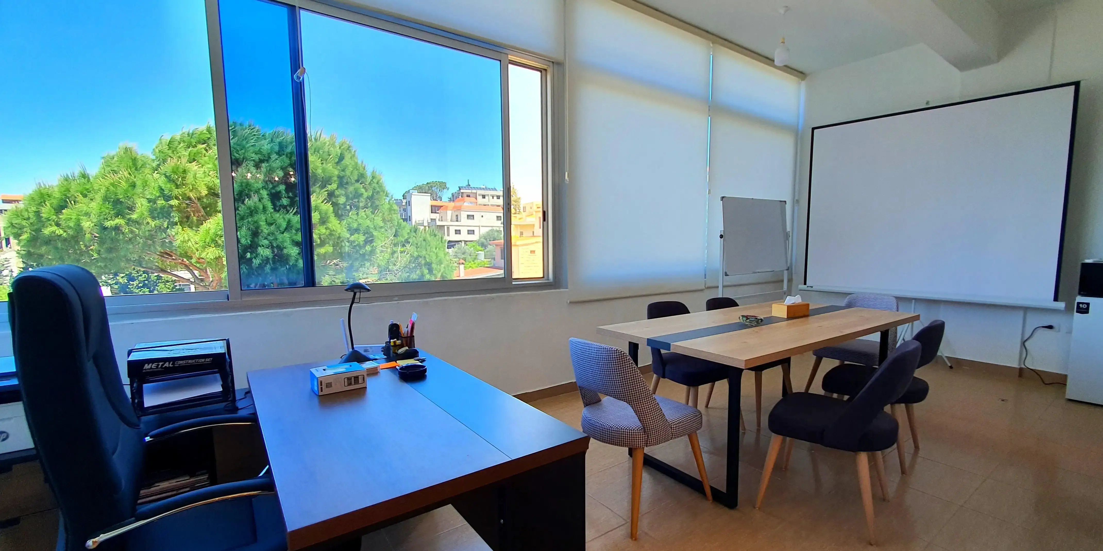 Training and workshops room in the clinic, showing a large table in the center, with a projector screen and whiteboard behind. Left to the table is an open view of the outside nature